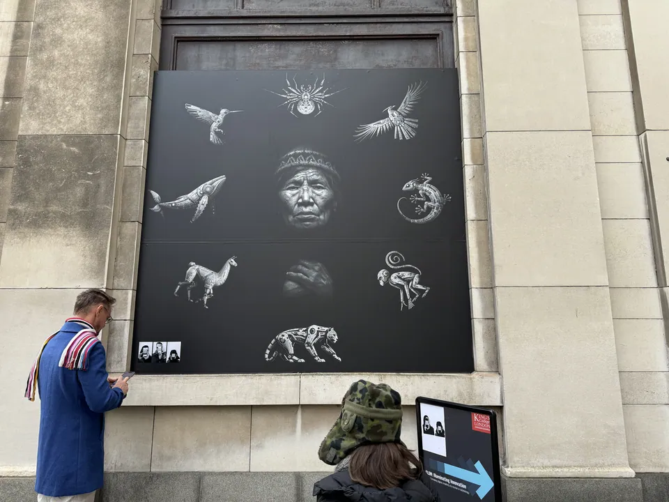 A large black panel of silver animals around a grandmother's face, on a stone building. A man looks at it.
