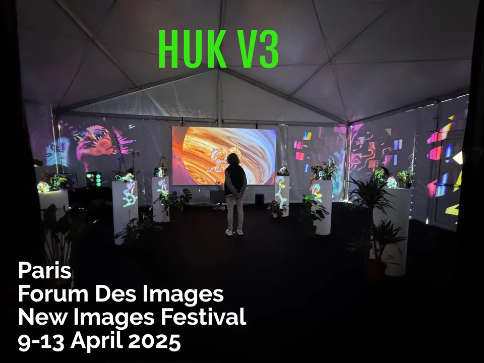 Visitantes en una carpa oscura de proyecciones, flores y pantallas. Arriba, las palabras Huk V3.