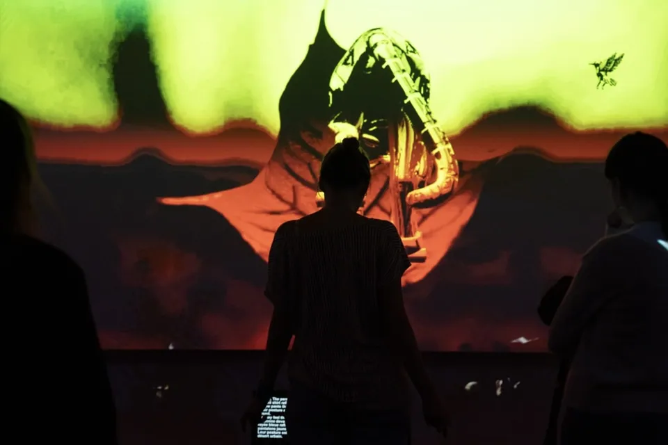 Visitantes en silueta frente a la proyección de una figura de jaguar contra un cielo rojo y verde.