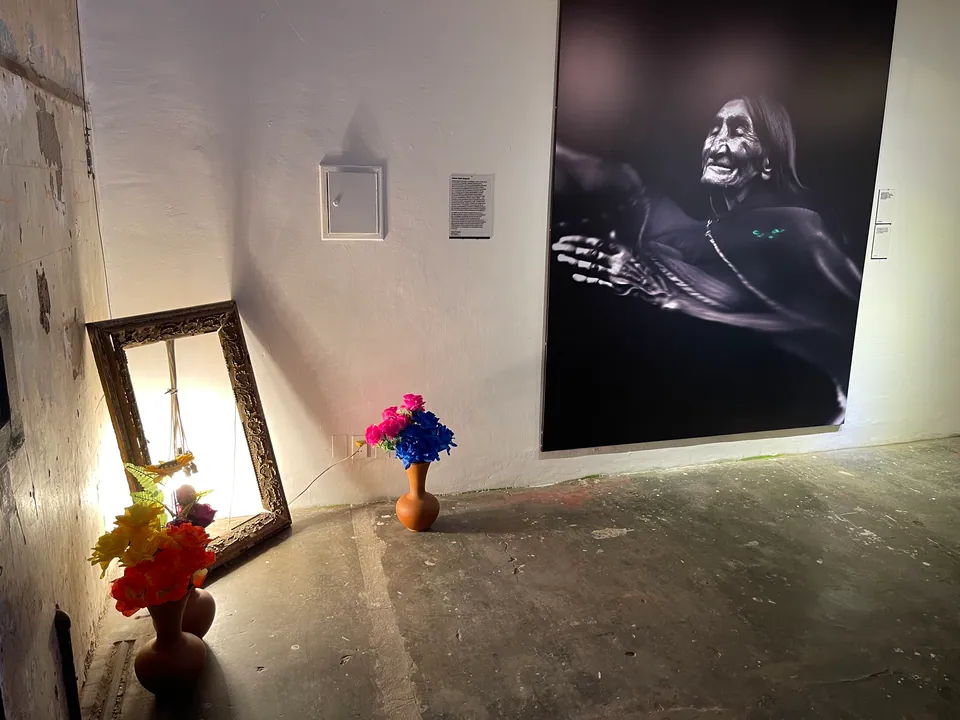 A large portrait of an old woman hangs above a mirror and vases of flowers on a cement floor.