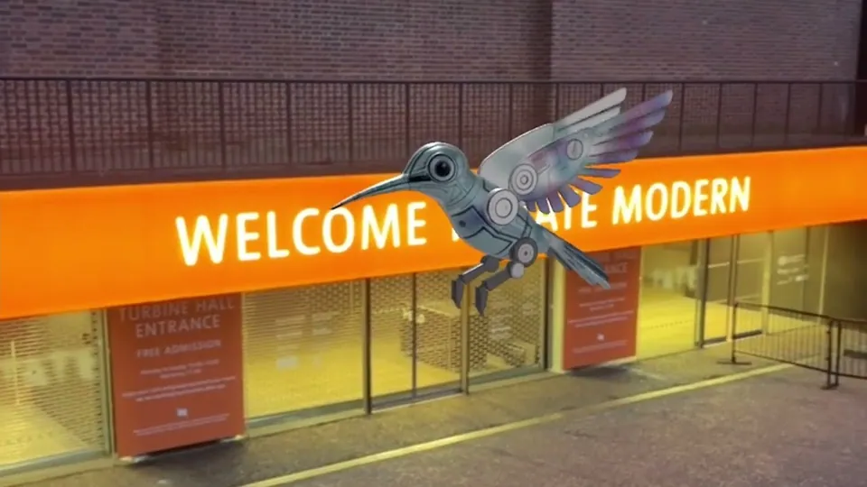 A silver robotic hummingbird flies in front of the entrance of Tate Modern.