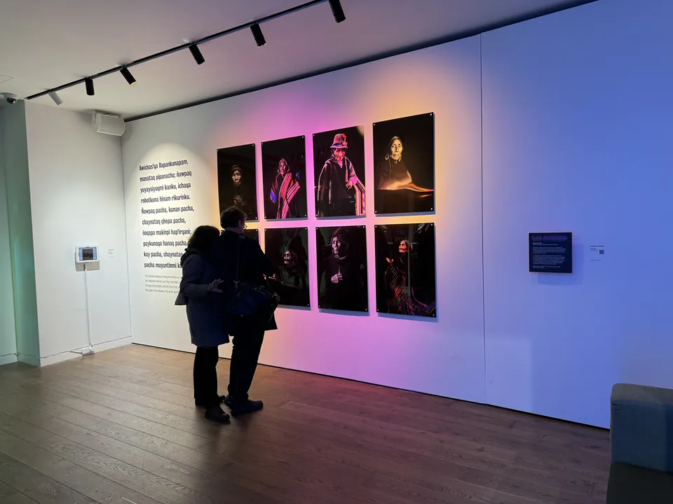 Two visitors stand before a wall of eight portraits lit in pink and blue.