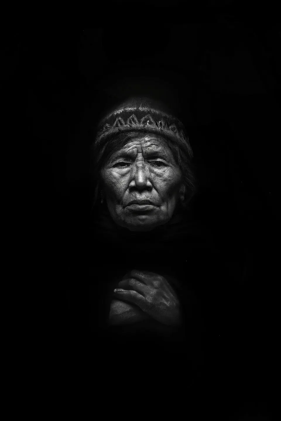 In black and white, a grandmother in a knitted hat looks out of the dark, her hands crossed on her chest.
