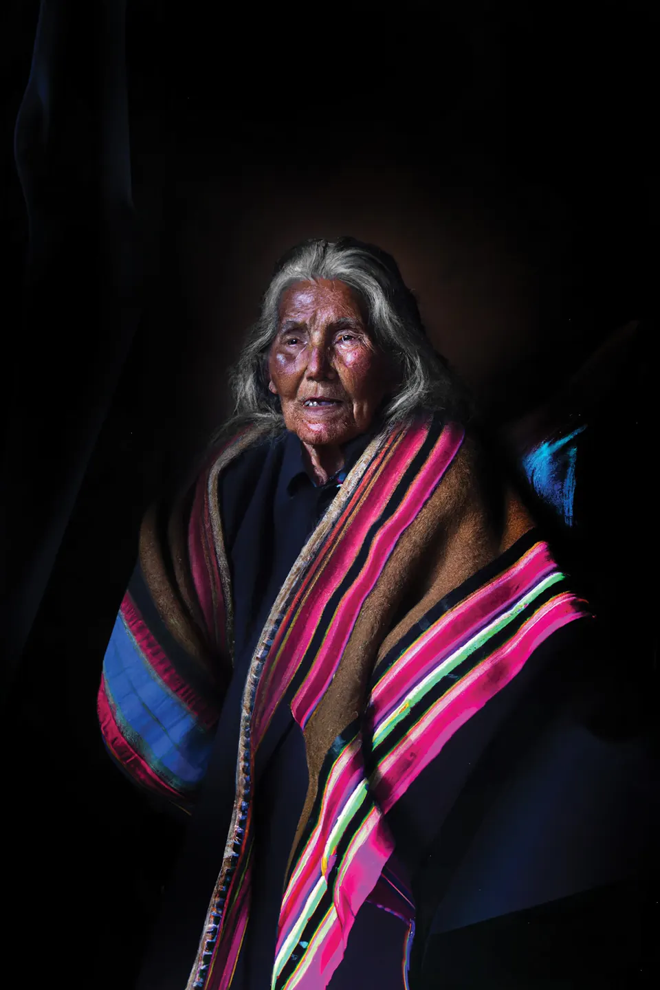 A grandmother with long grey hair wears a shawl striped in pink, blue and green, against black.