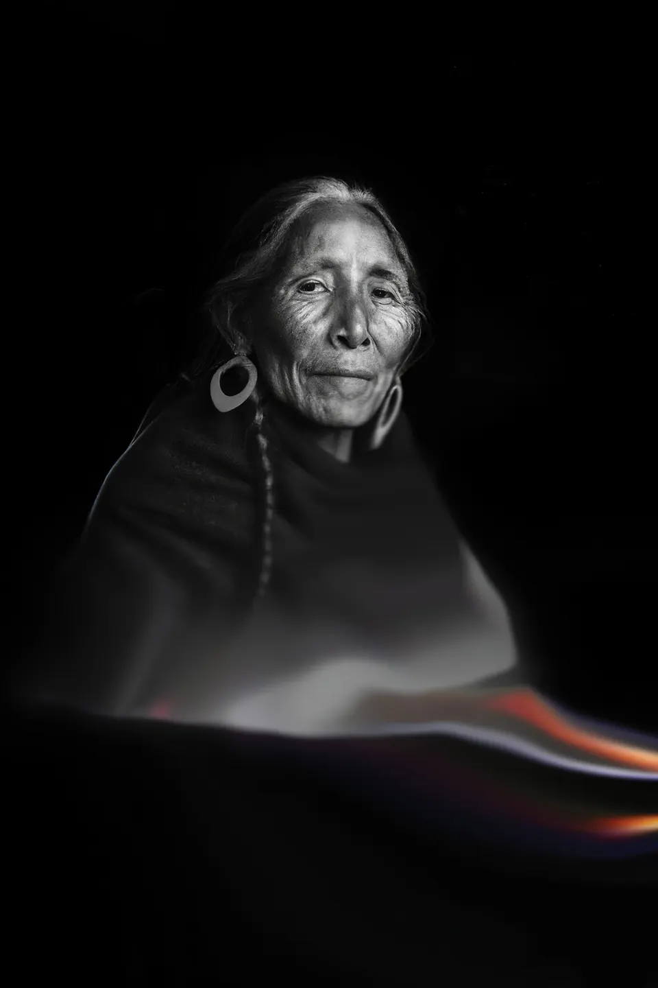 In black and white, a grandmother with a braid and large earrings, her shawl dissolving into a streak of colour.