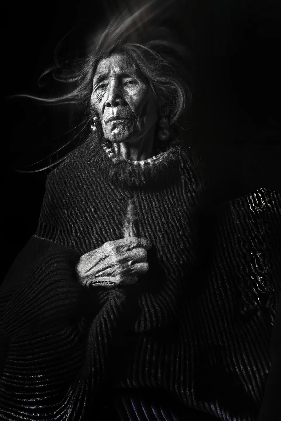 In black and white, a grandmother with windblown hair holds her hand to her chest, wrapped in a knitted shawl.