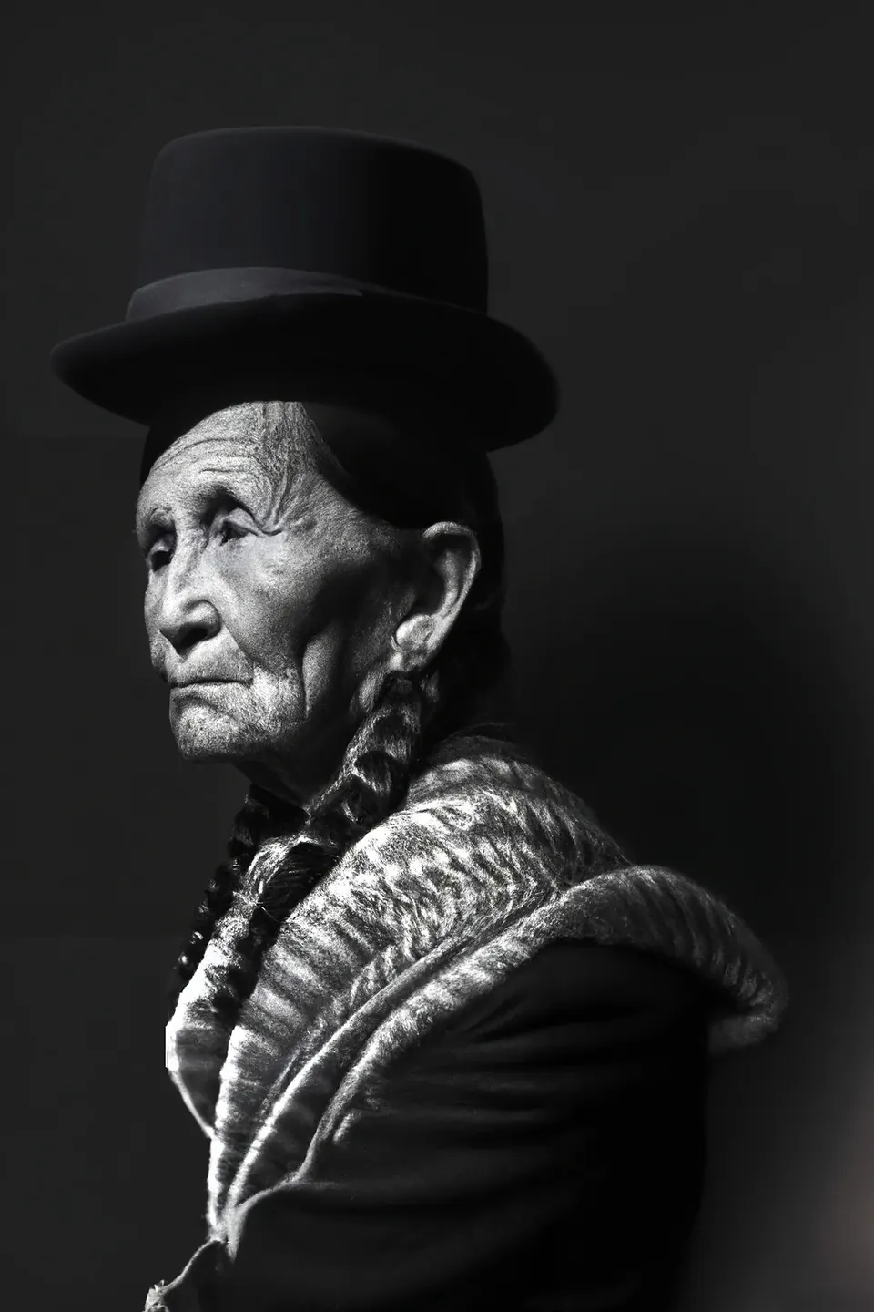 In black and white, a grandmother in a bowler hat, with long braids and a patterned shawl, seen in profile.