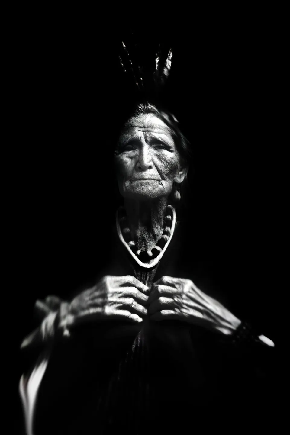 In black and white, a grandmother with a feather in her hair and a beaded necklace holds her hands at her chest.