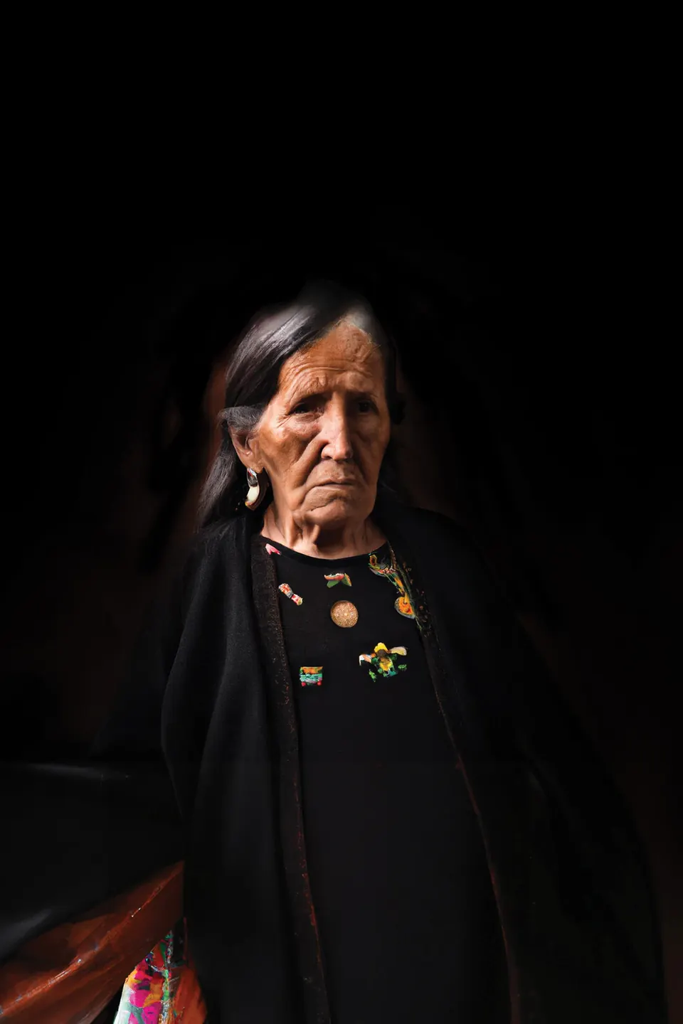 A grandmother in black, with small coloured embroidery, stands in the dark.