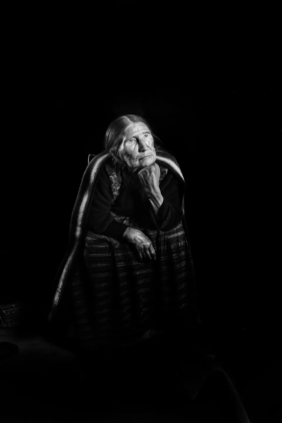 In the dark, a small grandmother sits hunched in a striped skirt under a single light.