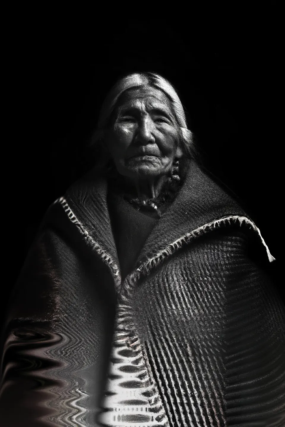 In black and white, a grandmother under a woven shawl whose stripes ripple like water.