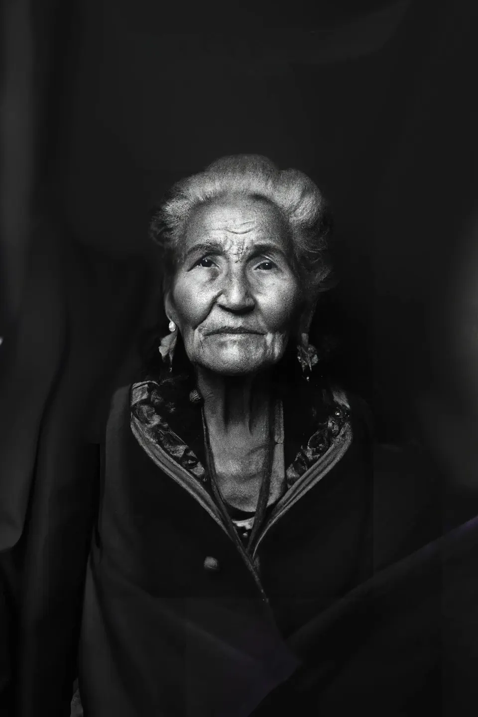 In black and white, a grandmother with white hair, earrings and an embroidered jacket.