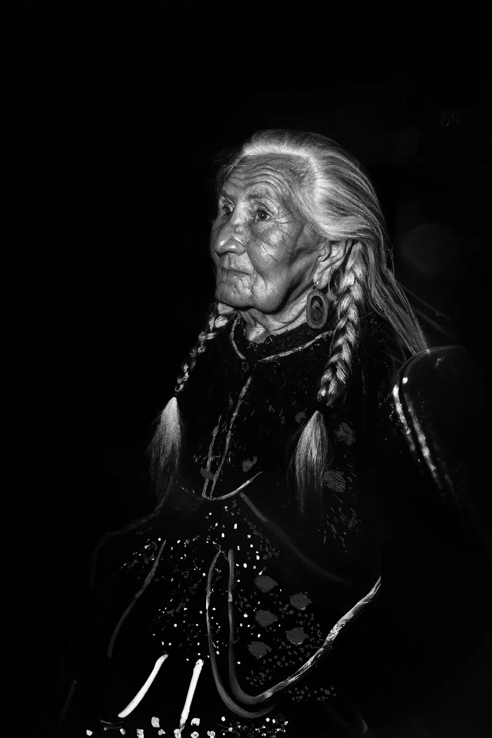 In black and white, a grandmother with long braids and a dark embroidered dress looks up.