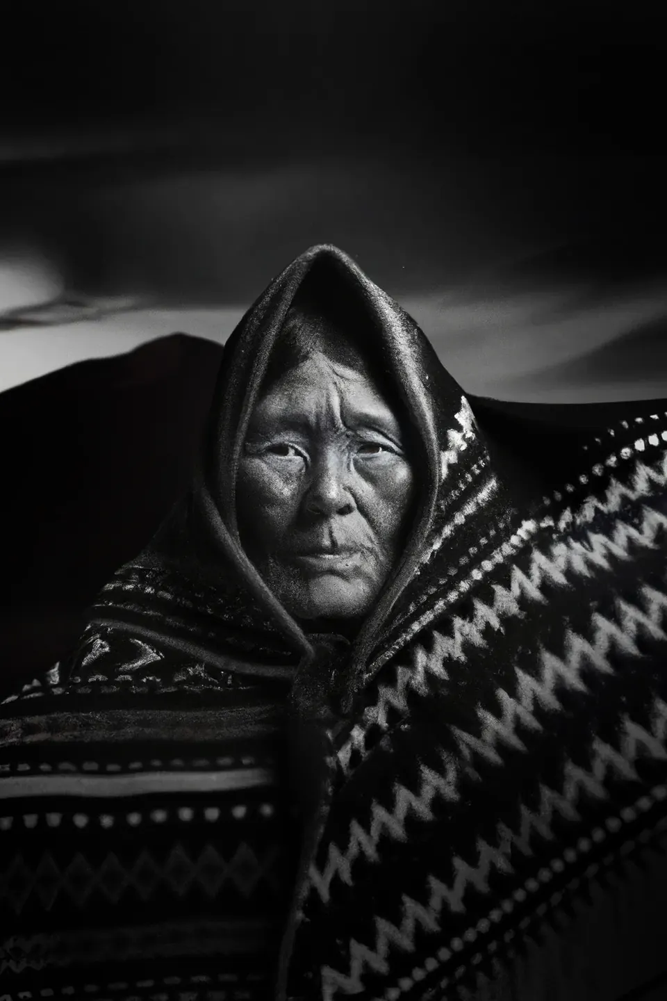 In black and white, a grandmother in a hooded zigzag shawl before dark mountains.