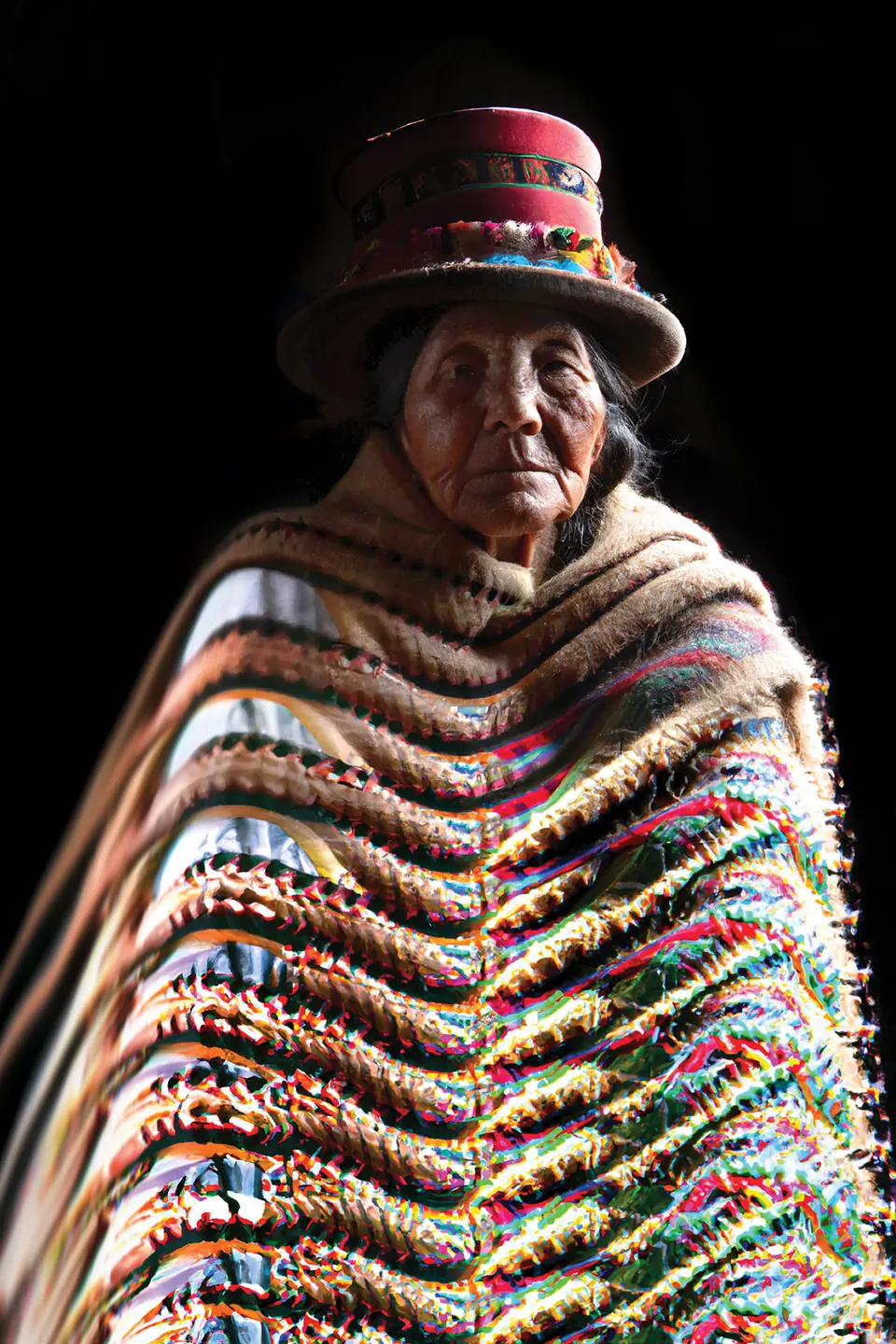 A grandmother in a red hat wears a fringed shawl of many coloured stripes.