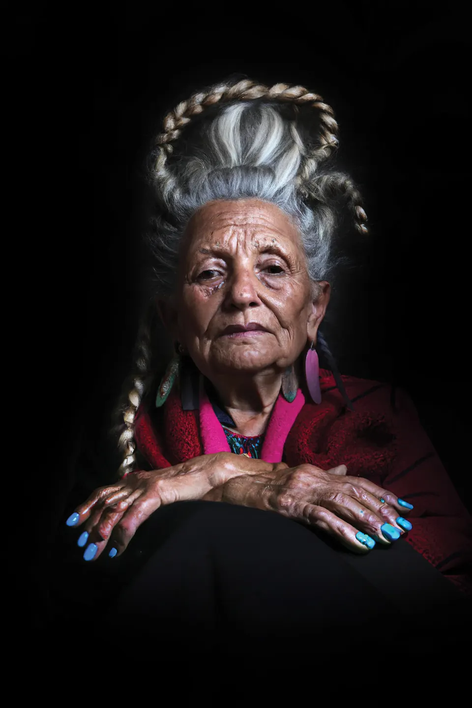 A grandmother with white braids piled high, blue nails and a red shawl, her hands on her knee.