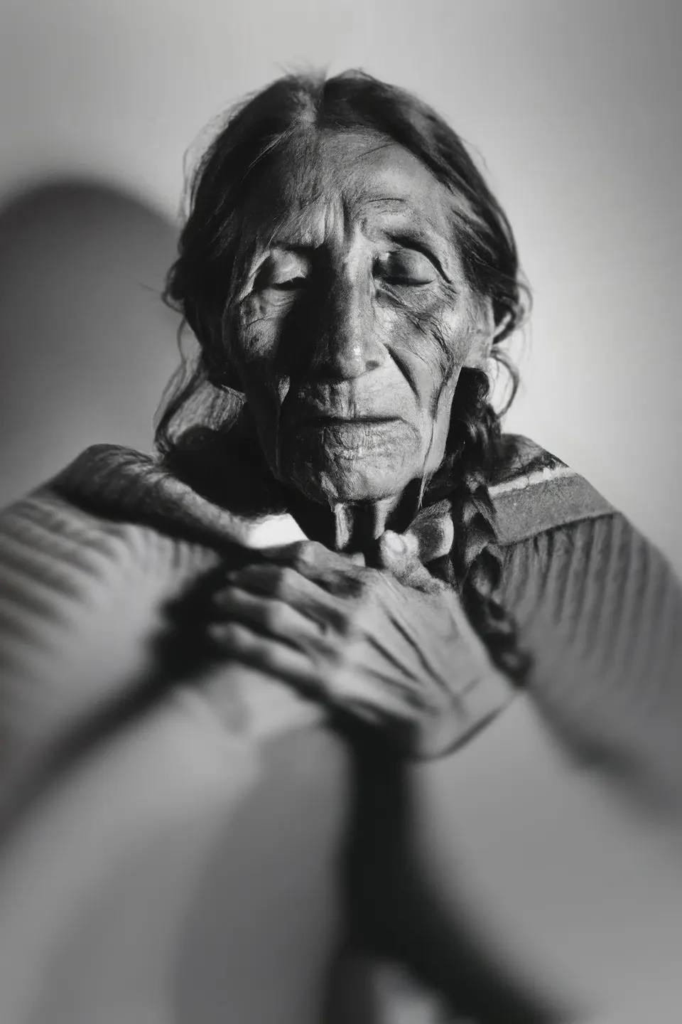 In black and white, a grandmother with braids and closed eyes holds her hands together.