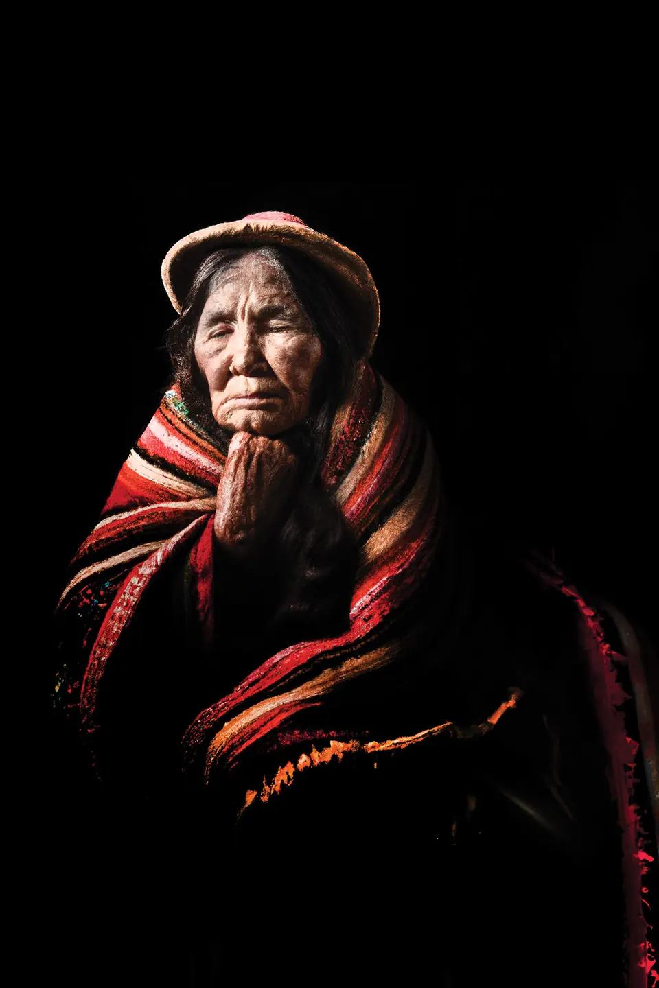 A grandmother in a straw hat rests her chin on her hand, wrapped in a red and gold striped shawl.
