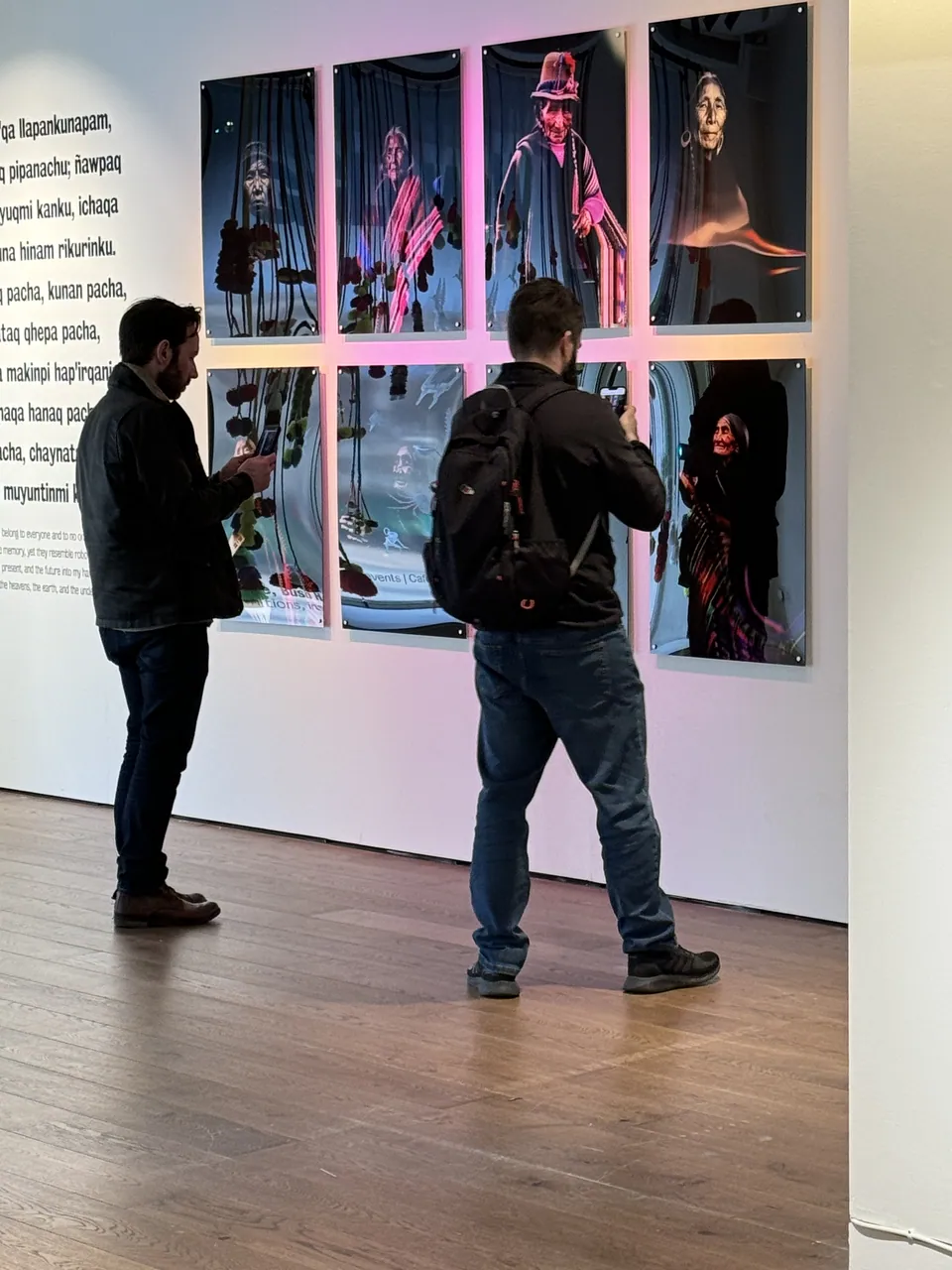 Two visitors read a wall of portraits lit in pink and violet.