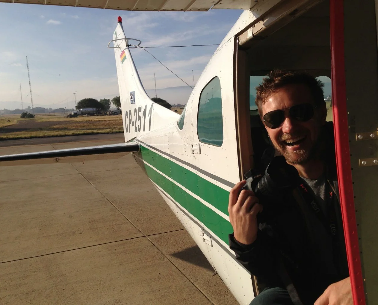 Un hombre con lentes de sol ríe en la puerta abierta de una avioneta verde y blanca.