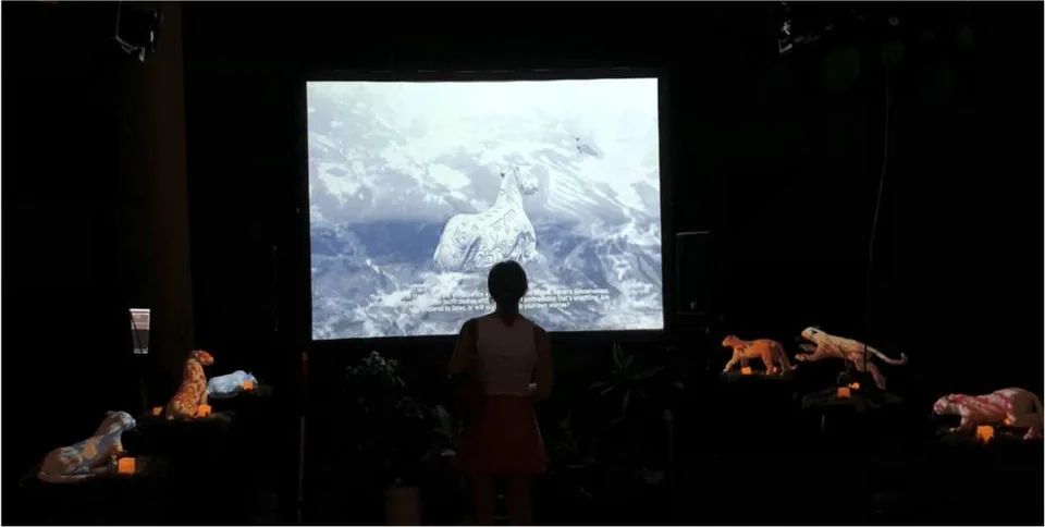 Una sala oscura: una persona de pie frente a una pantalla con un jaguar blanco, y jaguares pequeños iluminados en pedestales.