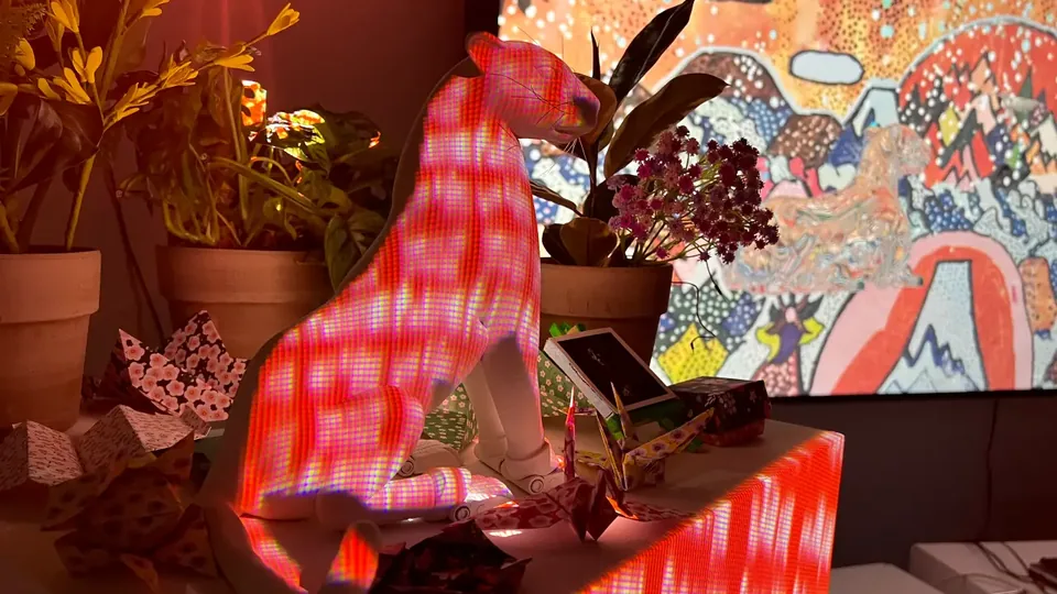 Una escultura de jaguar blanco iluminada con franjas rosadas y rojas, entre plantas y origamis, frente a un paisaje pintado.