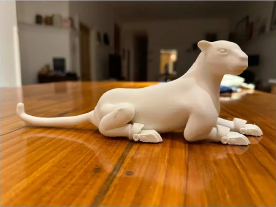 Un jaguar blanco impreso en 3D, con patas articuladas, echado sobre una mesa de madera.