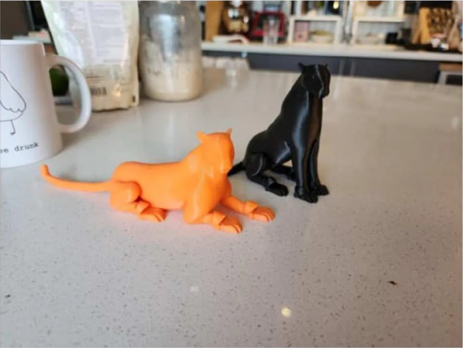Dos jaguares pequeños impresos en 3D sobre un mesón de cocina, uno naranja echado y uno negro sentado.