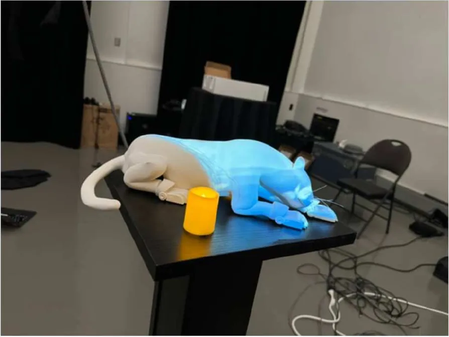Un jaguar blanco impreso, echado sobre un pedestal negro, iluminado de azul por un proyector, junto a un vasito amarillo.