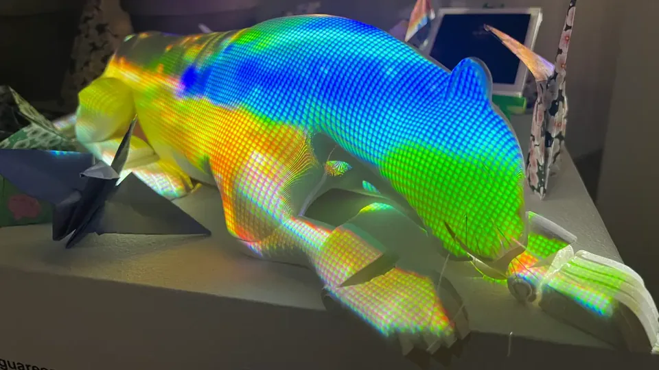 Un jaguar blanco echado, cubierto por una grilla de arcoíris proyectada, junto a grullas de papel.