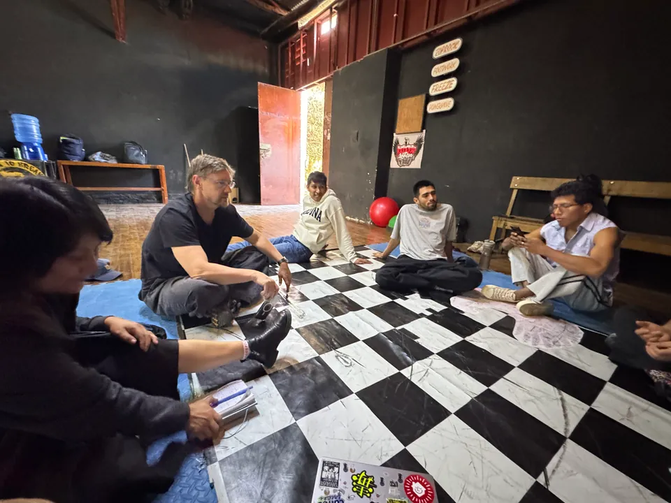 El equipo conversa en ronda en el piso a cuadros, alrededor de una laptop y hojas de papel.