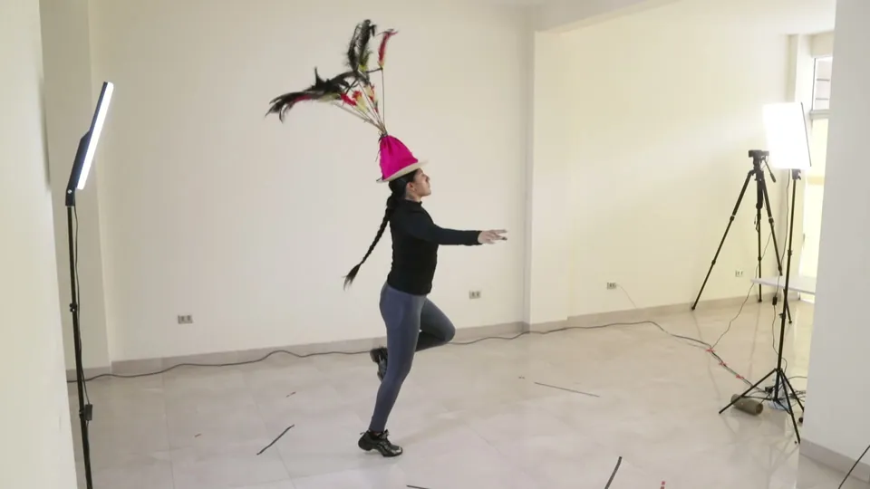 Una persona con un tocado alto de plumas gira sobre un pie en una sala blanca.