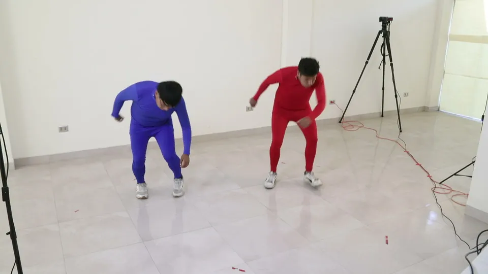 Dos bailarines con trajes azul y rojo bailan tinku, inclinados hacia adelante, en una sala blanca.