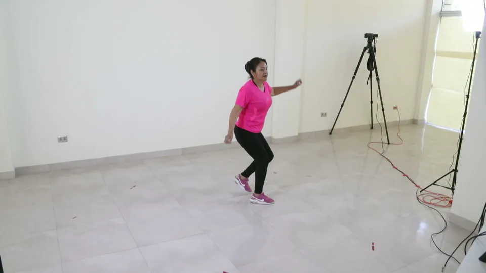 Una persona con polera rosada baila sobre un piso blanco, junto a un trípode.