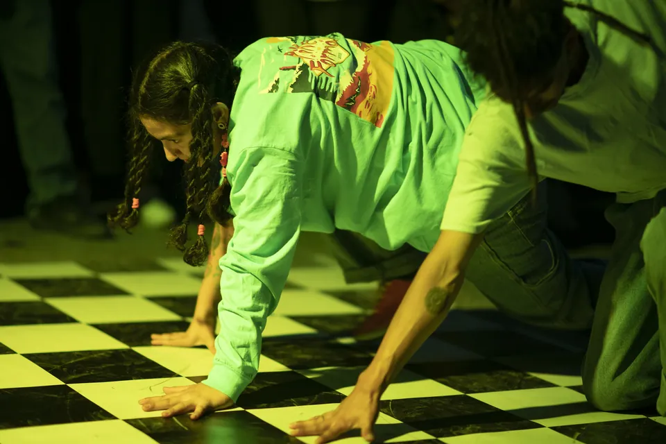 Una bailarina gatea sobre un piso a cuadros iluminado de verde, junto a otro bailarín.