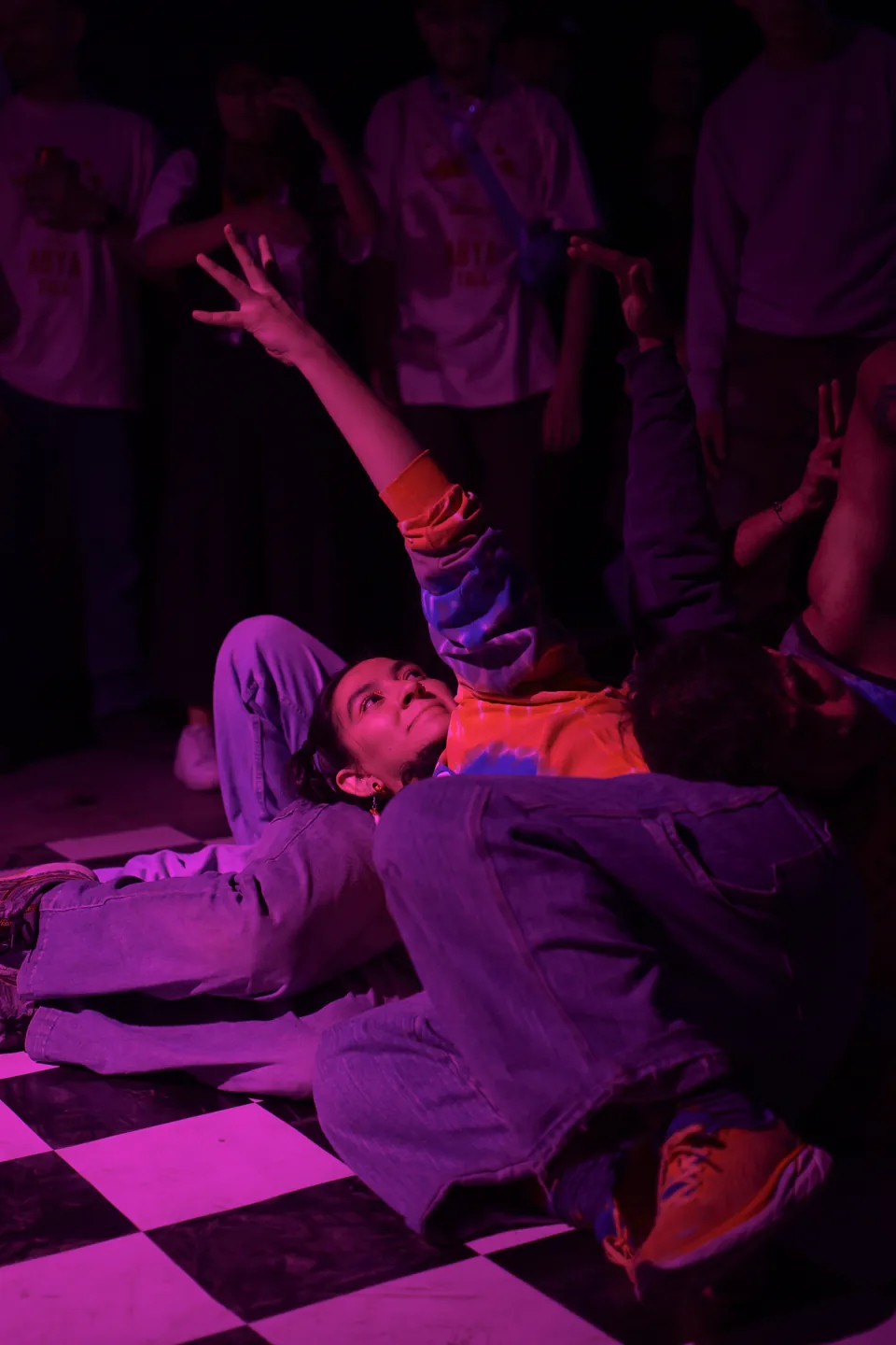 Una bailarina en el piso estira el brazo hacia arriba bajo luz magenta.