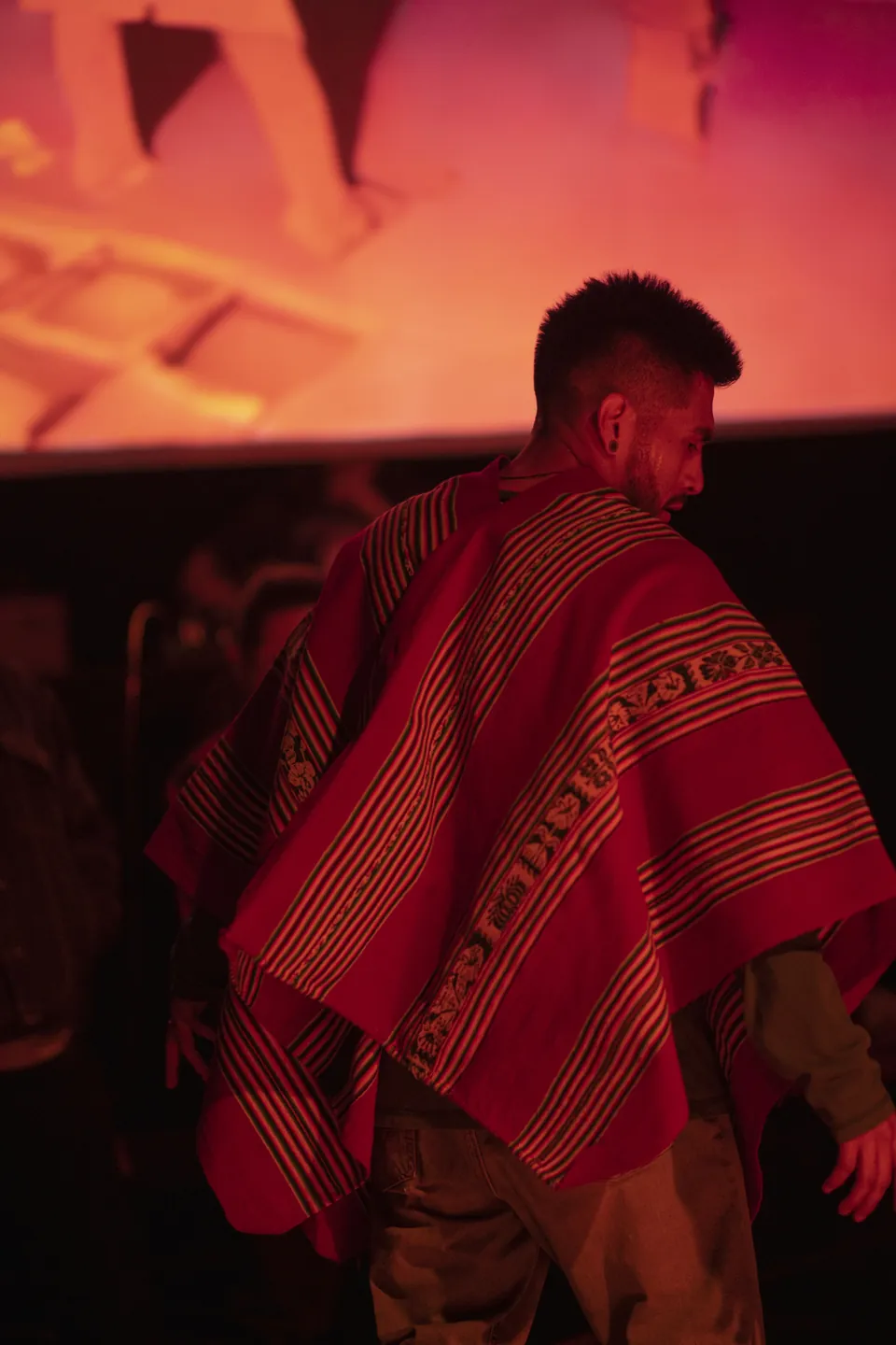 Un bailarín con un poncho rojo a rayas, de espaldas, bajo luz roja.