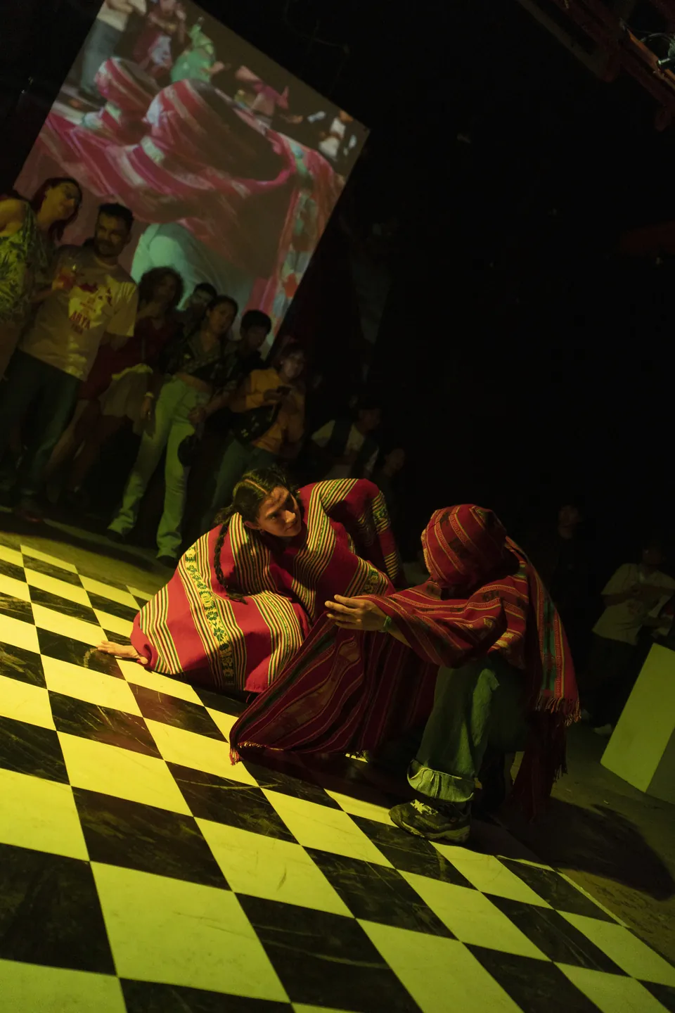 Bailarines envueltos en ponchos rojos, agachados sobre un piso a cuadros amarillo.