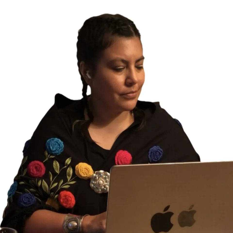 Una mujer con trenzas y un chal negro bordado con flores trabaja en una laptop.