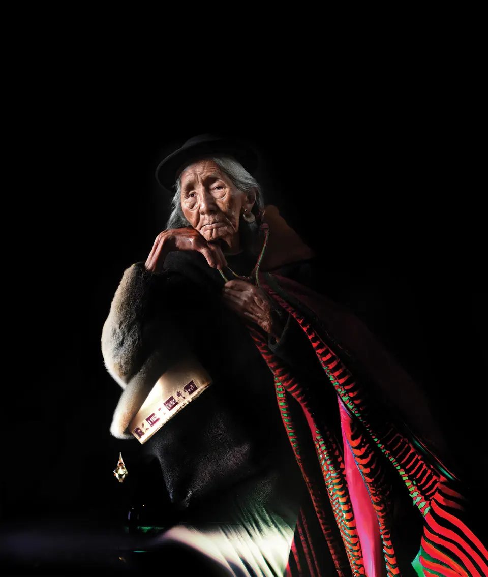 Un retrato de Las Awichas: una mujer mayor con sombrero oscuro apoya el mentón en la mano. Su manta se vuelve franjas rojas y verdes.