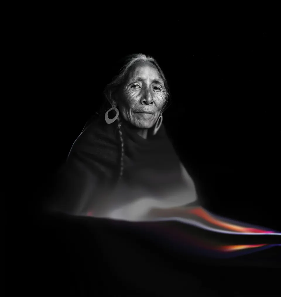 Un retrato de Las Awichas en blanco y negro: una mujer mayor con una trenza y un arete grande. Una banda de color pasa debajo de ella.