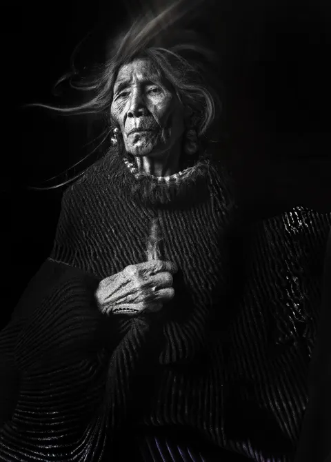 Un retrato de Las Awichas en blanco y negro: una mujer mayor con el pelo al viento se lleva una mano al pecho.