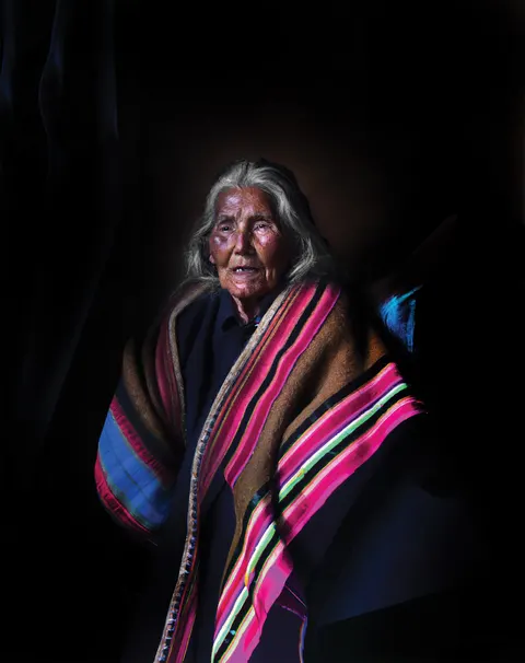 Un retrato de Las Awichas: una mujer mayor de pelo canoso lleva una manta de franjas rosadas, azules y verdes.
