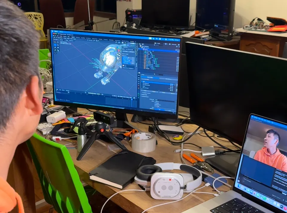 A person sits at a desk in front of a monitor that shows a 3D creature in Blender. A laptop shows a video call. A VR headset lies on the desk.