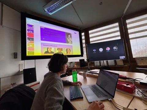 A person works on a laptop in a meeting room. A large screen shows a page that reads Feminist AI Residents.