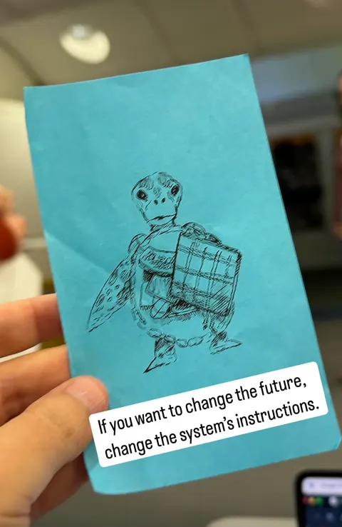 A hand holds a blue card with a drawing of a turtle that carries a bundle. A caption reads: If you want to change the future, change the system's instructions.
