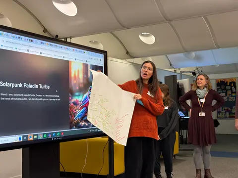 A person in a red jumper holds up a large sheet of notes beside a screen that reads Solarpunk Paladin Turtle. Another person stands behind.