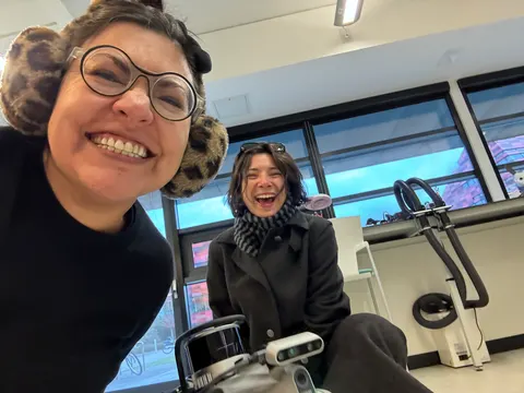 Two people laugh at the camera in an office. One wears leopard print earmuffs and round glasses.