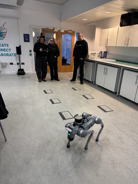 A four-legged robot stands on the floor of a lab. Three people watch from the back of the room.