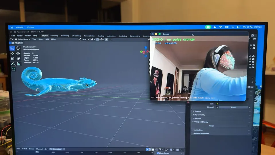 A monitor shows a turquoise gecko model in Blender. A small window shows a person with headphones and tracking points on the face.